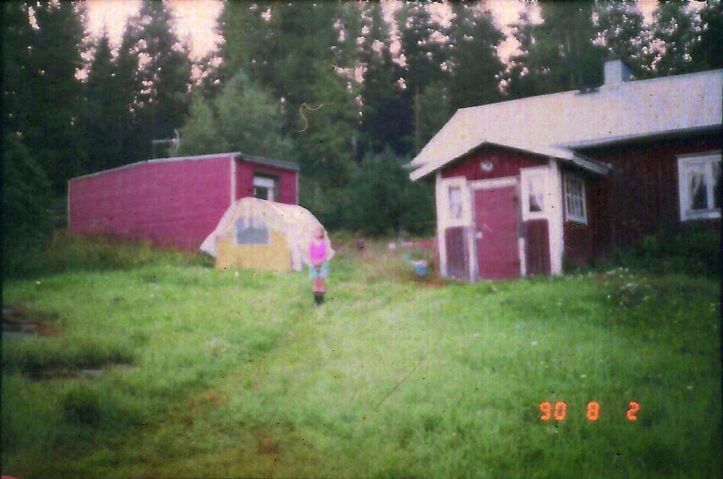 A blurred photo of a child standing in front of a cottage, a mobile home and a tent, surrounded by green grass and with the forest in the background. The photo's burned-in date stamp is 90 8 2.