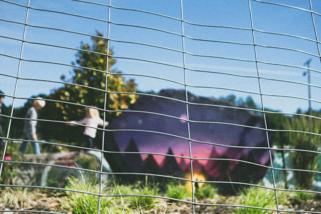 a close up on wire fencing in the foreground with children playing outdoors in the background and the image of a twilight pink and purple sky with a bright light and a silhouette in the center of the light