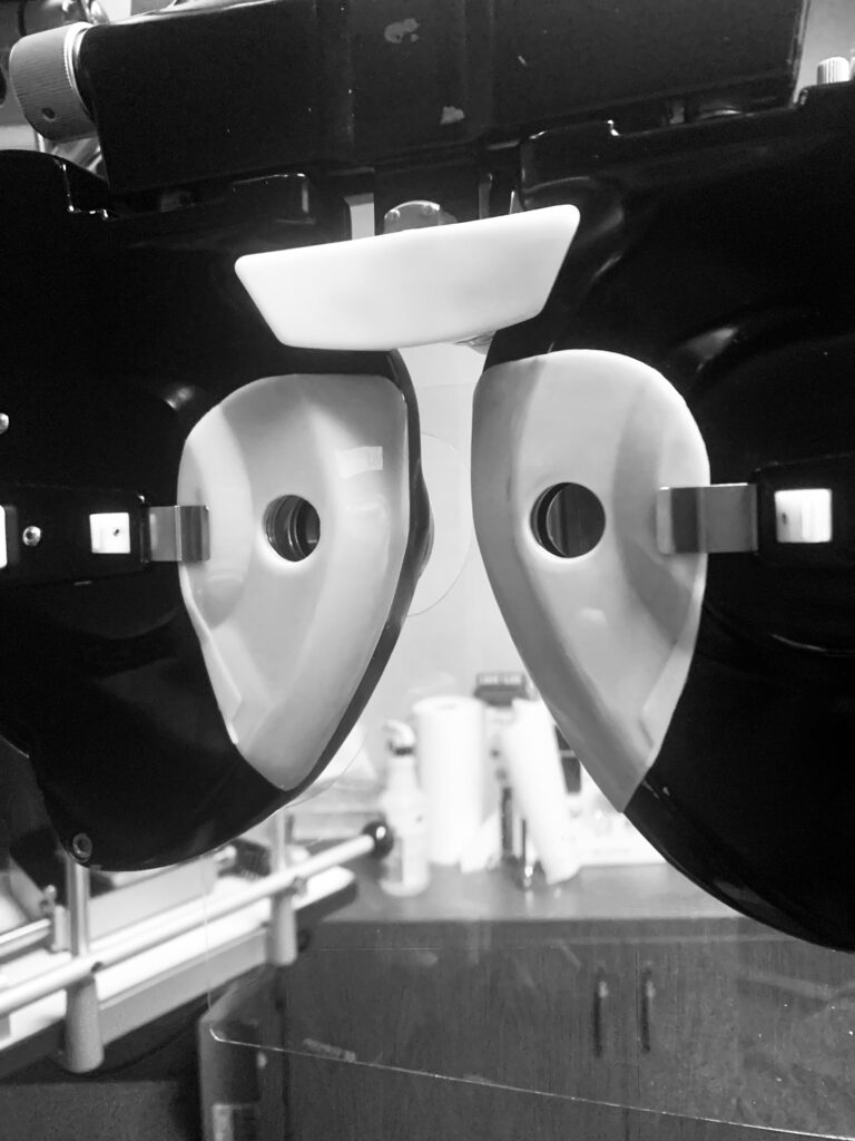 "A? Or B?" is a black and white photo taken in the office of an optometrist from the perspective of the patient chair. The opthalmoscope takes up the upper 3/4 of the photograph and appears close to the lens, as if it is about to be pushed towards the patient's face. You see only the parts of the machine that come up towards the face, and the eyeholes appear at the center of the photograph. The machine appears in full focus. You can see through the gaps in it and under the machine in depth-blur a further part of the optometrist's office, with a cabinet with rolls of paper towels and spray bottle on it, conceivably to clean the opthalmoscope between patients.



This photo asks us to examine the concepts of clarity, truth, and integrity. What in the photo is the way we are supposed to see it? Once we are using the opthalmoscope, which eye sees with integrity with which lens. A? Or B? Sometimes, it is hard if not impossible to discern. 