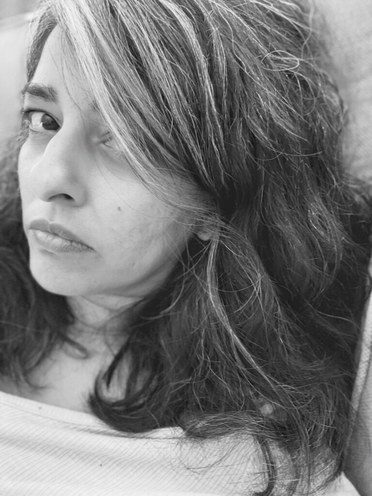 Migraine Trigger, MD
Black and white portrait photograph of middle-aged, South Asian woman looking into camera. Upper half of the shot frames her face. Top right of photo is darker toned, with her cascading hair, and bottom left of photo is lighter-toned with her face, neck and top of shirt. Her hair is black and wavy with grey streaks that frame her face. Her left eye is partially covered by her hair, while her right eye is fully exposed and conveys a knowing, a pleading, or maybe even a resignation. She looks utterly fed up. She sits upright, resting her head back against a cushion. Her lips are pursed into a slight frown. There is an intimacy to this photo, with the subject connecting directly with the viewer with vulnerability we usually shy away from in polite society. The subject appears too exhausted to care about politeness or masking, and her expression and posture convey a quiet, seething rage. This work is called, "Migraine Trigger, MD," and is from a series of self-photographs titled, "Portraits of Pain + Peace," capturing the artist trying to live, survive, and cope with all within their body and all outside of it. This piece was shot after returning from a medical visit in which the artist once again was gaslit, disbelieved, and dismissed, and thus made more sick by the system claiming to make them less sick.