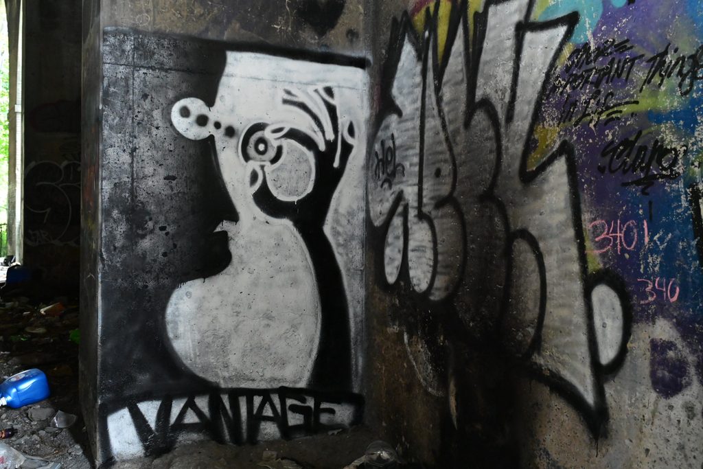 Photograph of graffiti on a wall. The photo is looking into a corner. On the left is the central image, a black and white drawing of a person plucking out its eye, signed "Vantage" below. The wall on the right has black and white graffiti lettering. 