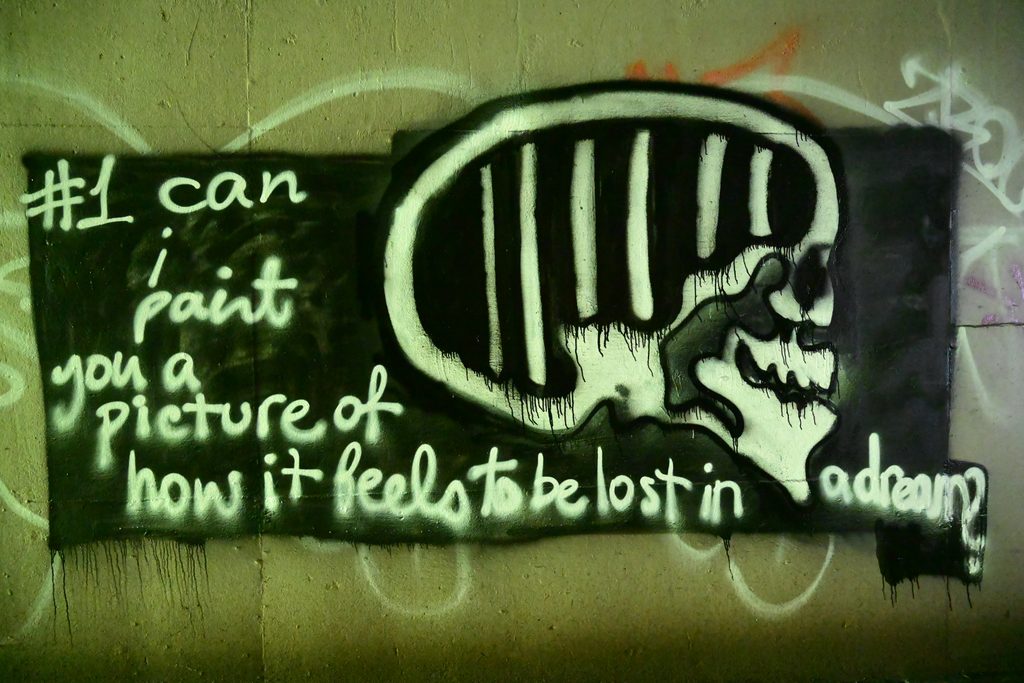 Graffiti drawing of a black rectangle with a skull and the words, "#1: can I paint you a picture of how it feels to be lost in a dream?" The edges of the rectangle and text are slightly blurred. The black rectangle is painted over a green wall with graffiti. 